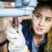 A person wearing gloves is fixing a leaking pipe with a wrench. Water is spraying from the pipe. The focus is on the hands and the pipe, with the person's face blurred in the background.