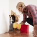 A woman kneeling on the floor, using a sponge to clean up water. There is a red bucket nearby. The wall in the background is damaged, showing pipes behind it.