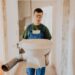 A person wearing safety goggles and blue overalls is carrying a detached toilet in an unfinished room. The walls are bare, showing exposed concrete, and there are some bricks stacked in the background. The individual is holding the toilet with gloved hands.